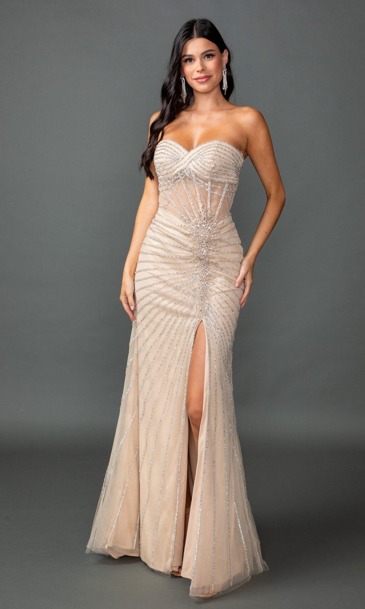 A woman stands against a plain gray background wearing the Dancing Queen 4562 long prom dress in champagne, featuring a strapless, fitted silhouette with silver embellishments, a corset bodice, and a thigh-high slit.